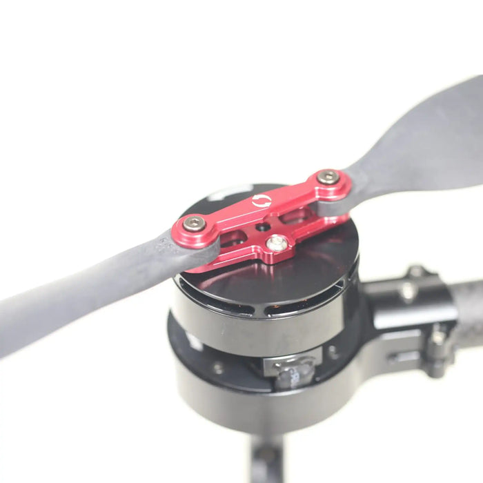 Close-up of a red anodized aluminum propeller blade attachment bracket mounted on a black motor hub, with a gray propeller blade extending into the frame, set against a clean white background.