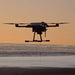 A black AEE Condor Pro Fishing drone flying over a beach at sunset, with its payload suspended by a thin cable below it.