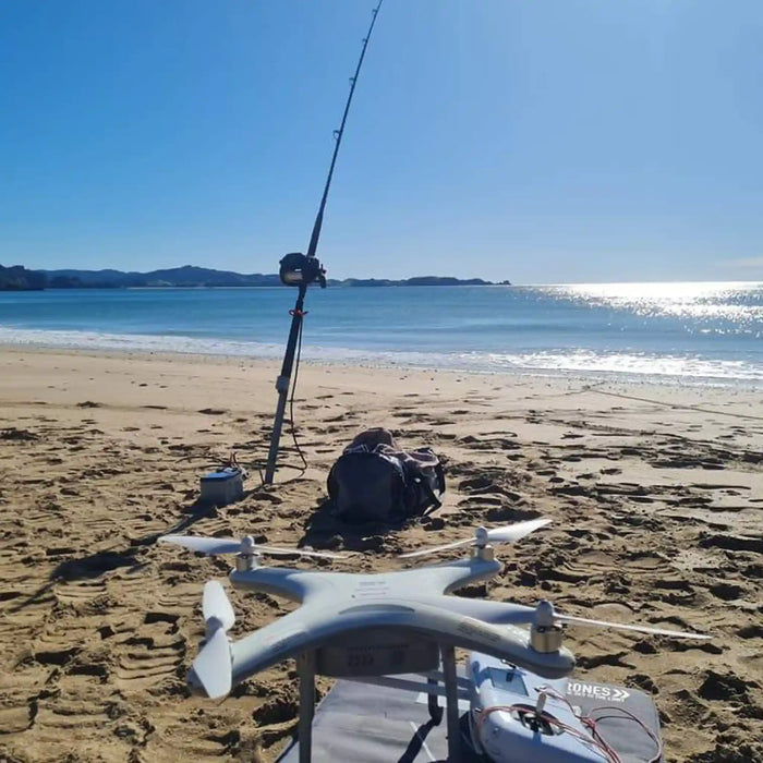 A white Condor A22S Fishing drone sits on a beach next to a fishing rod on a stand, with the ocean and shoreline in the background under a clear blue sky.